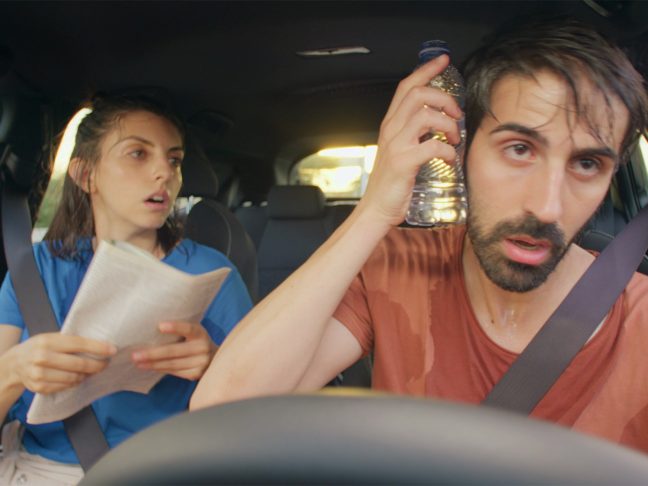 Man and woman in car sweating, trying to cool down by flagging paper and holding a water bottle